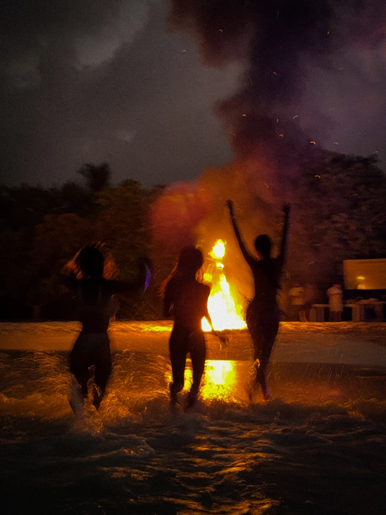 Three silhouettes running into the sea beside a beach fire at night