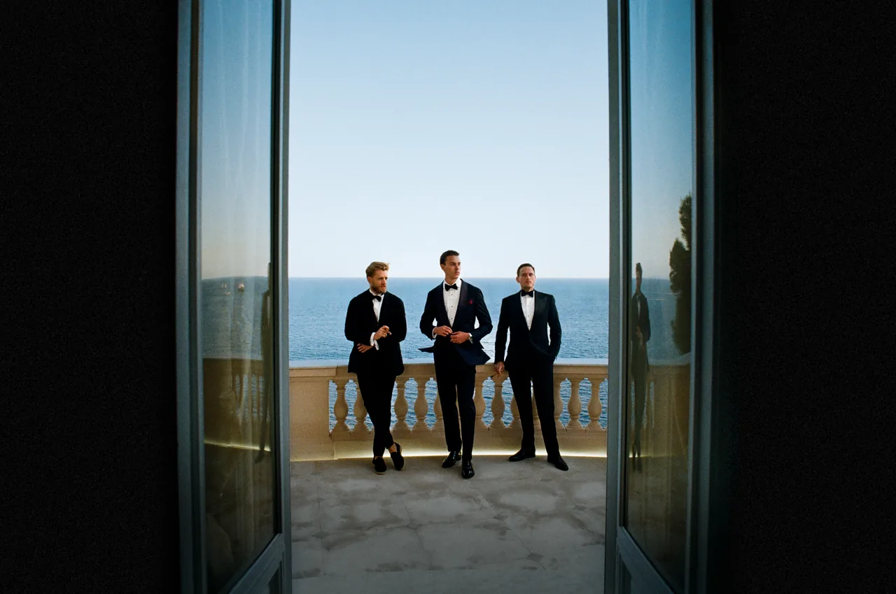 Three men in black tie on a balcony above the sea, framed by open doors