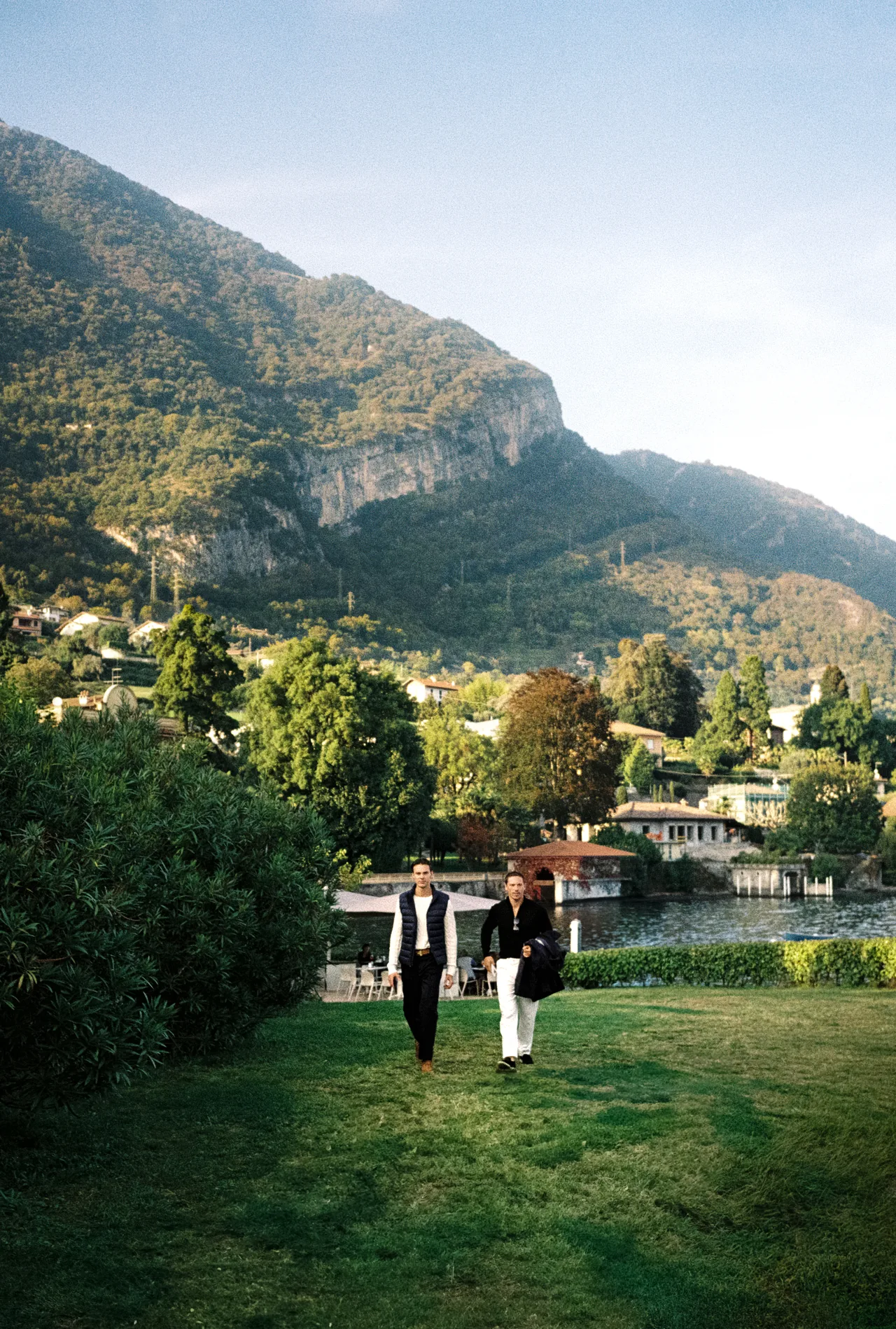 Two men walking across a lakeside lawn beneath a mountain